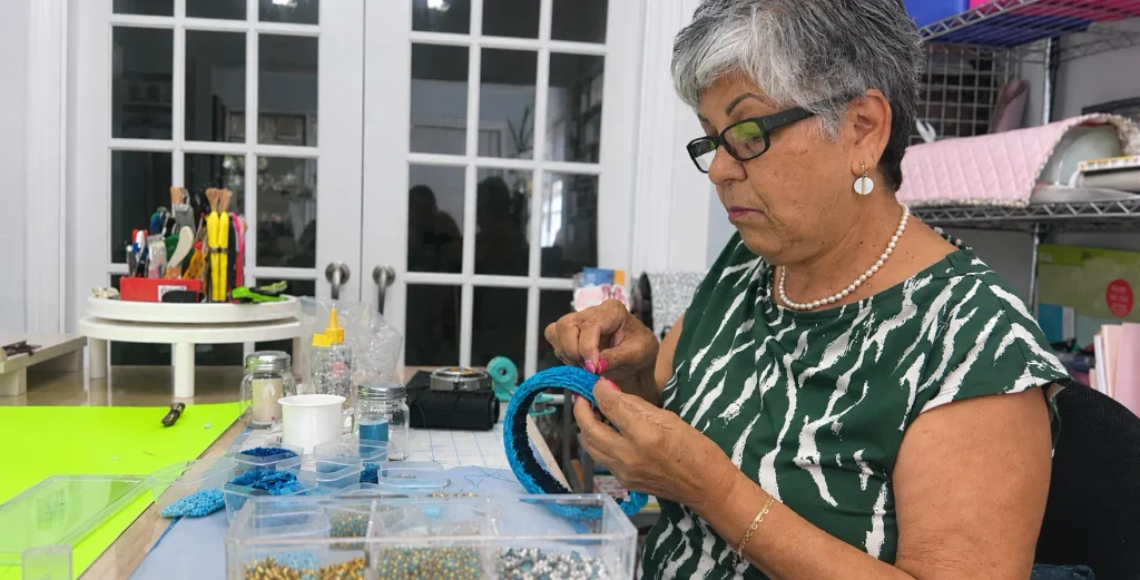 Woman creating a handmade headband by hand in a craft studio, carefully adding beads to a blue headband surrounded by beading supplies and tools.